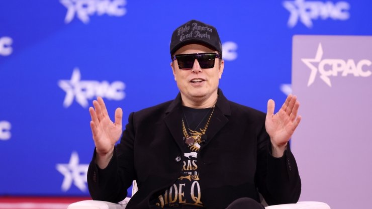 Elon Musk sitting and holding his hands up on stage at the 2025 Conservative Political Action Conference